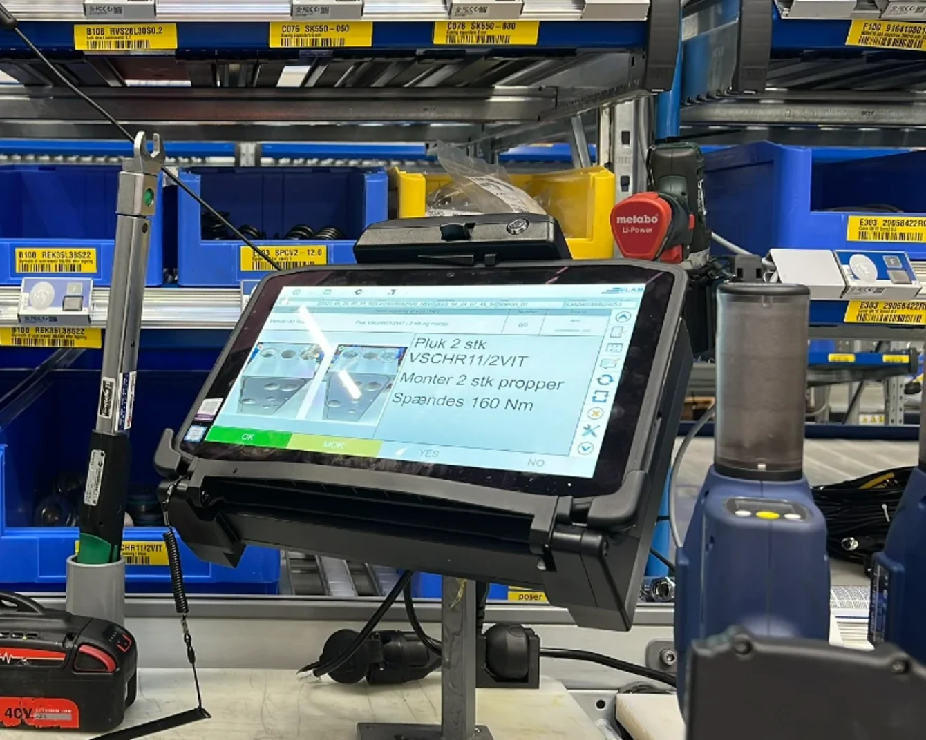 Tablet på et stativ viser instruktioner i et lager med blå kasser og værktøj i baggrunden.