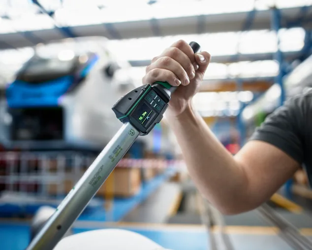 Persona usando una llave dinamométrica digital. El par es visible en una pequeña pantalla. El fondo desenfocado muestra un tren en una fábrica de construcción ferroviaria.
