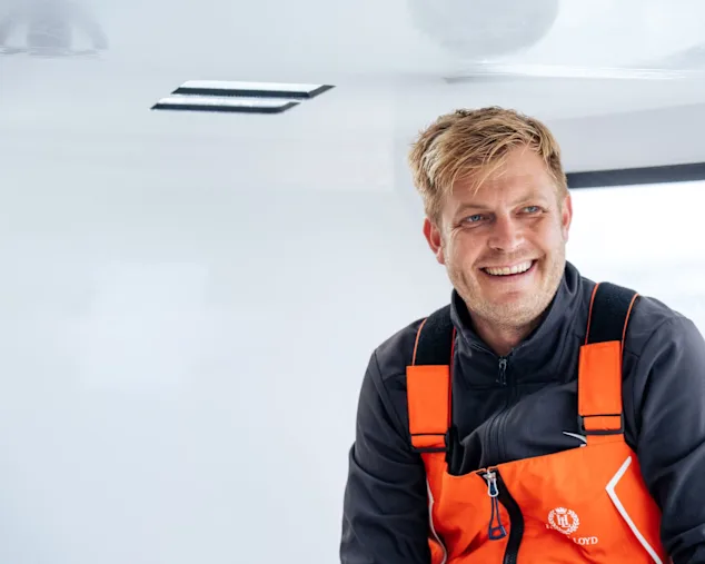 Glimlachende Oliver Heer in een oranje en zwart reddingsvest aan boord van een witte boot met ramen zichtbaar op de achtergrond.