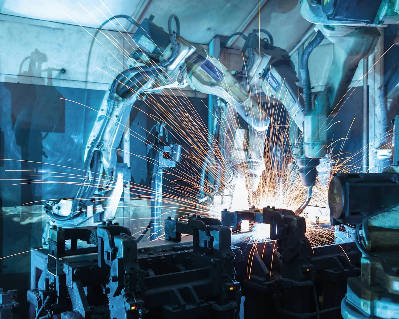 Przemysłowe ramiona robotyczne spawające metal z jasnymi pomarańczowymi iskrami w niebiesko oświetlonej fabryce