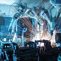 Przemysłowe ramiona robotyczne spawające metal z jasnymi pomarańczowymi iskrami w niebiesko oświetlonej fabryce