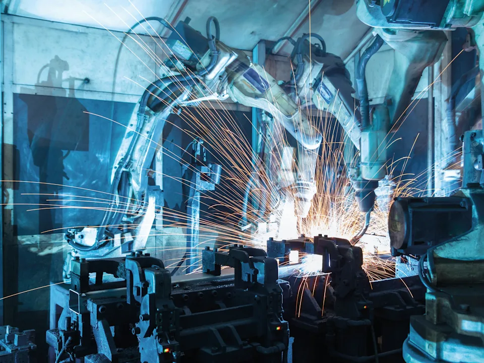 Industrial robotic arms welding metal with bright orange sparks flying in a blue-lit factory setting.