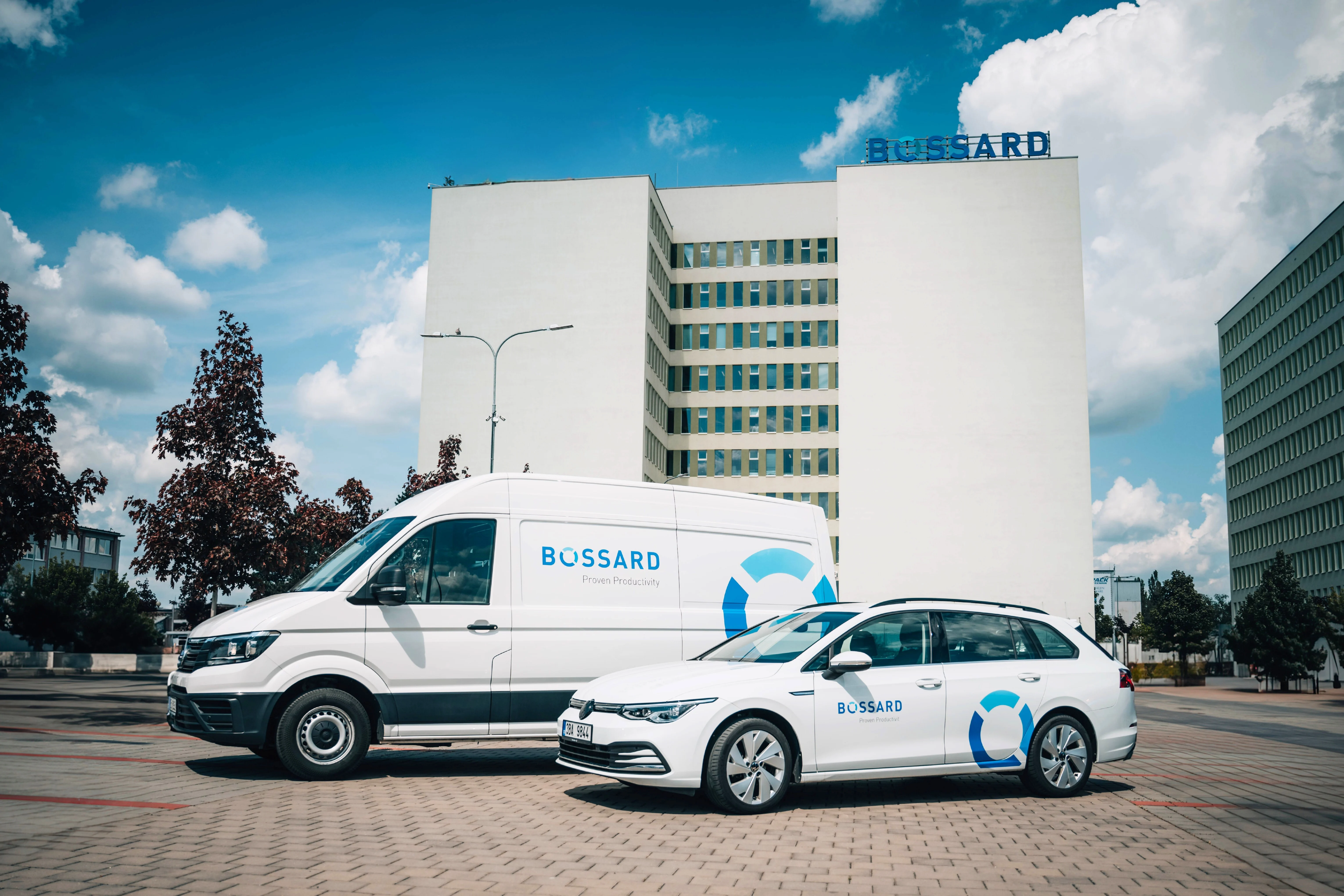 Dvě bílá vozidla s logy "Bossard" zaparkovaná před vysokou budovou pod modrou oblohou s mraky.