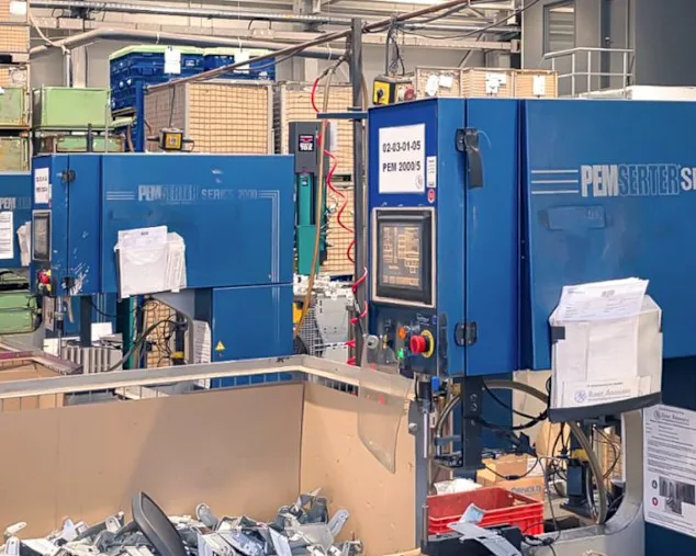 Zwei blaue PEMSERTER® Einpressmaschinen der Serie 2000 in einer Produktionshalle von Alzner Automotive, umgeben von Werkstücken, Kisten und Produktionsmaterialien.
