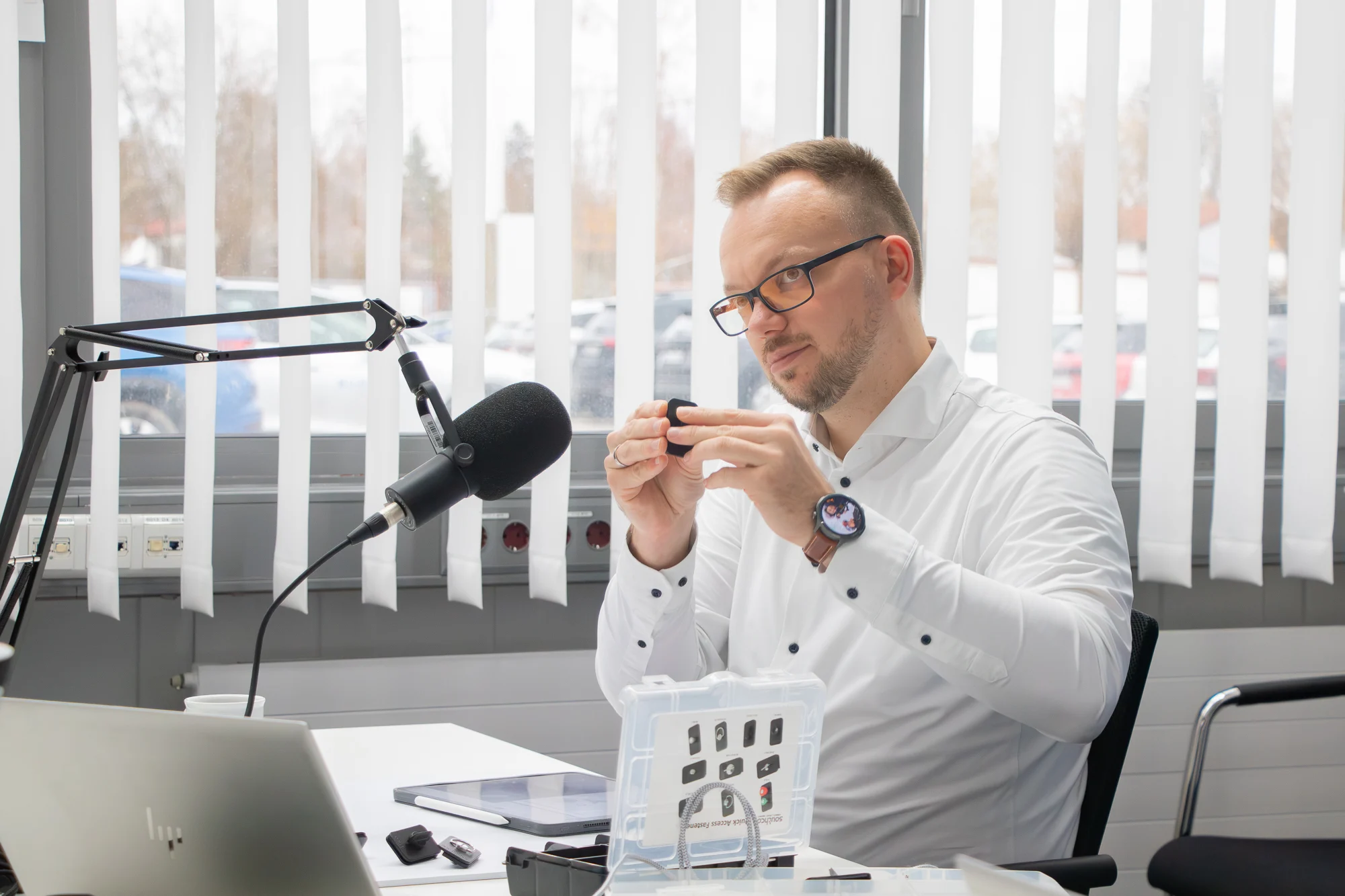 Tobias Engelberg in weißem Hemd sitzt in einem hellen Raum vor einem Broadcast Mikrofon. Er hält einen Vierteldrehverschluss vor sich hoch. Eine Musterbox, ein Tablet und Notebook stehen vor ihm auf dem Tisch.