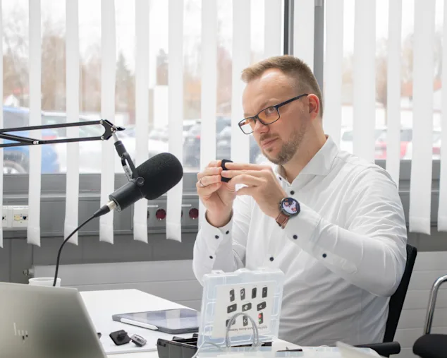 Tobias Engelberg in weißem Hemd sitzt in einem hellen Raum vor einem Broadcast Mikrofon. Er hält einen Vierteldrehverschluss vor sich hoch. Eine Musterbox, ein Tablet und Notebook stehen vor ihm auf dem Tisch.