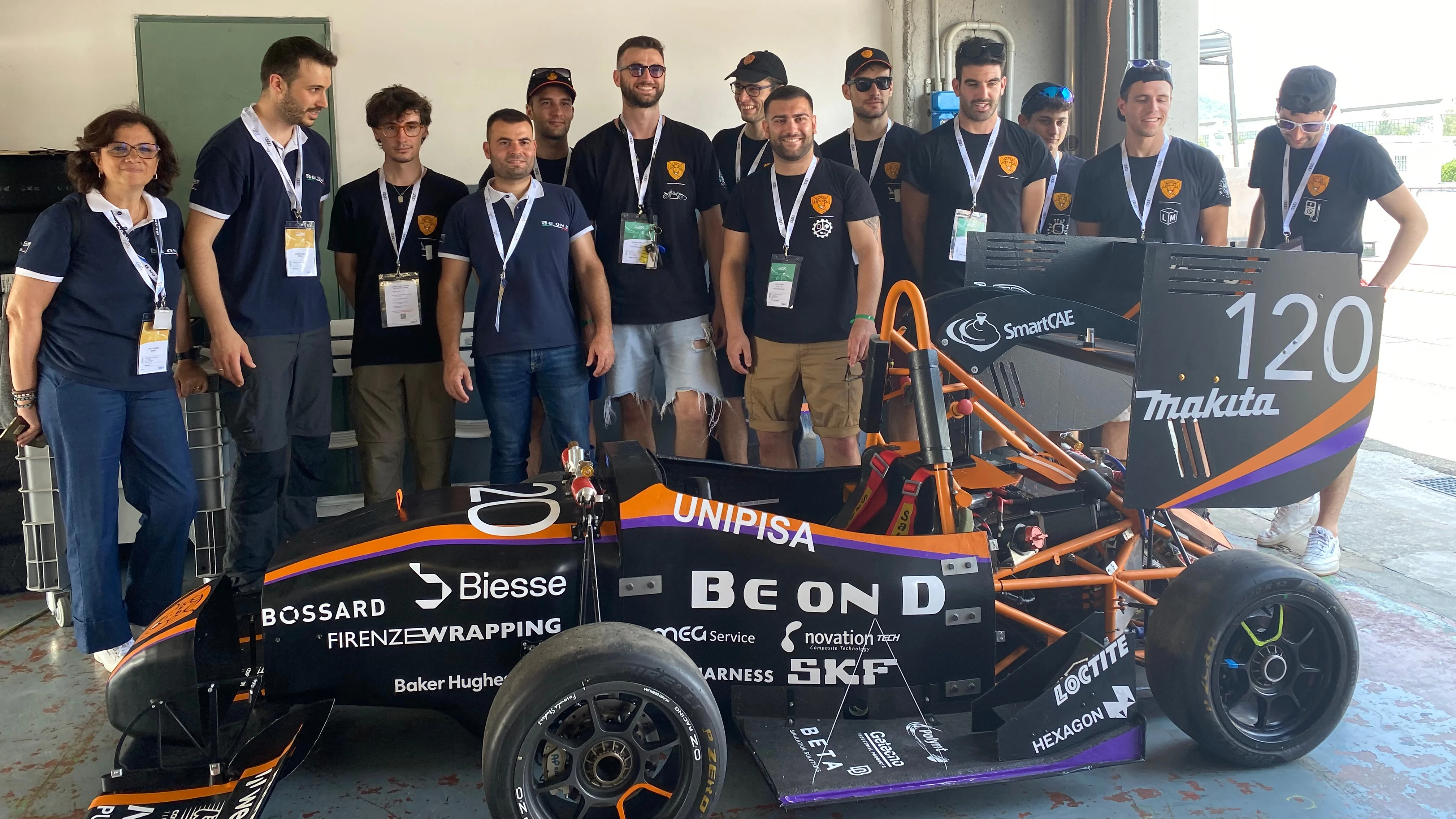 Un gruppo di persone con magliette abbinate si trova intorno a un'auto da corsa nera e arancione con il numero 120 e vari loghi di sponsor.