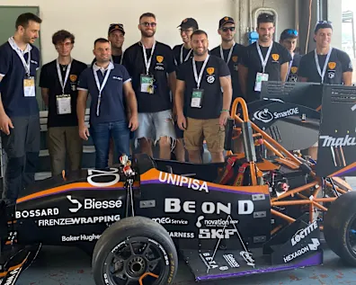 Un gruppo di persone con magliette abbinate si trova intorno a un'auto da corsa nera e arancione con il numero 120 e vari loghi di sponsor.