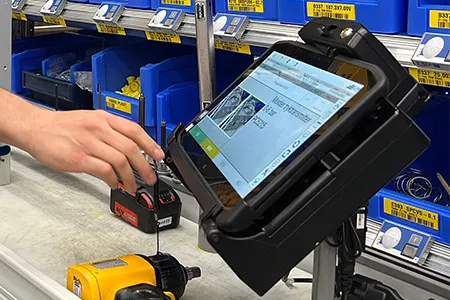 Una mano interagisce con un tablet su un supporto in un'officina, circondata da attrezzi e contenitori blu con etichette.