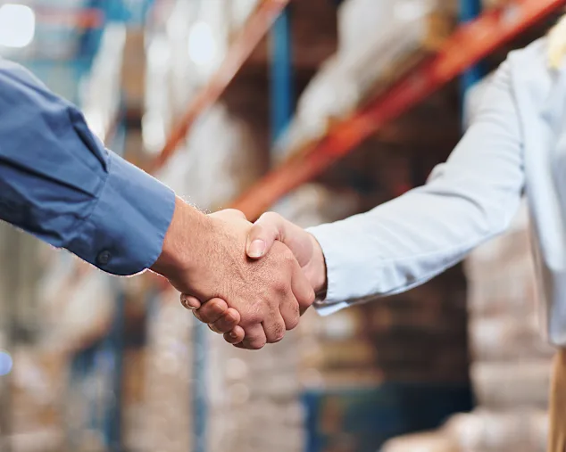 Two people shaking hands in a warehouse, one wearing a blue shirt and the other a white shirt with a name badge.