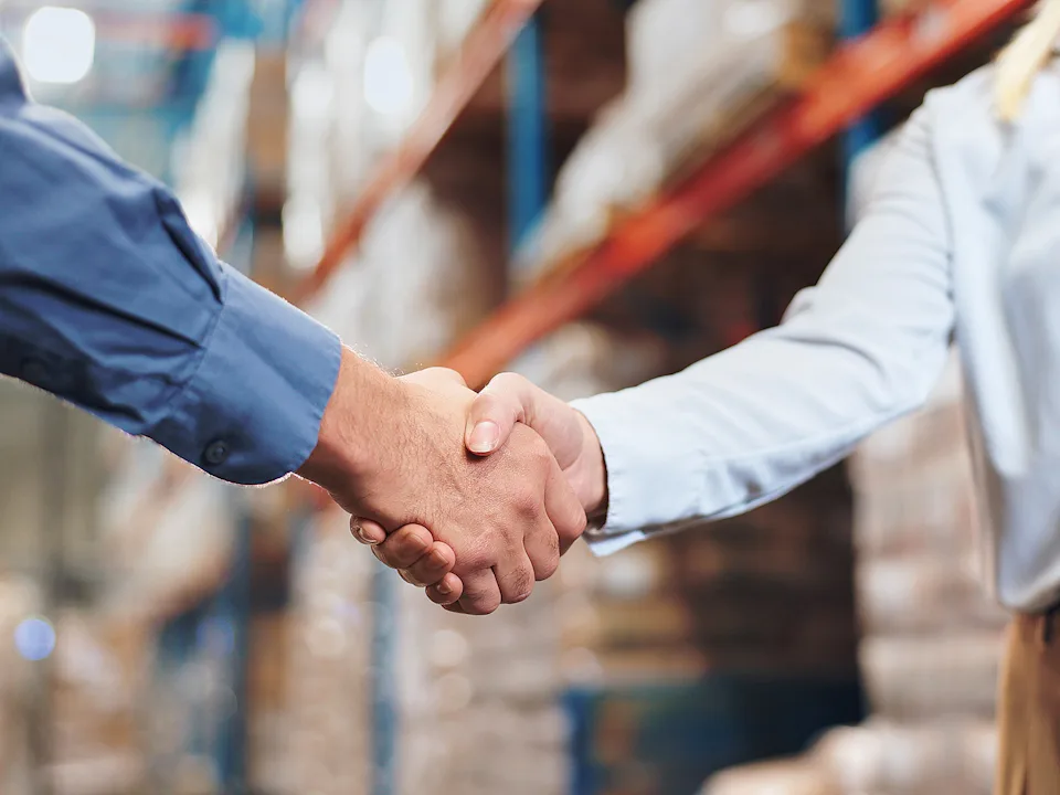 Two people shaking hands in a warehouse, one wearing a blue shirt and the other a white shirt with a name badge.