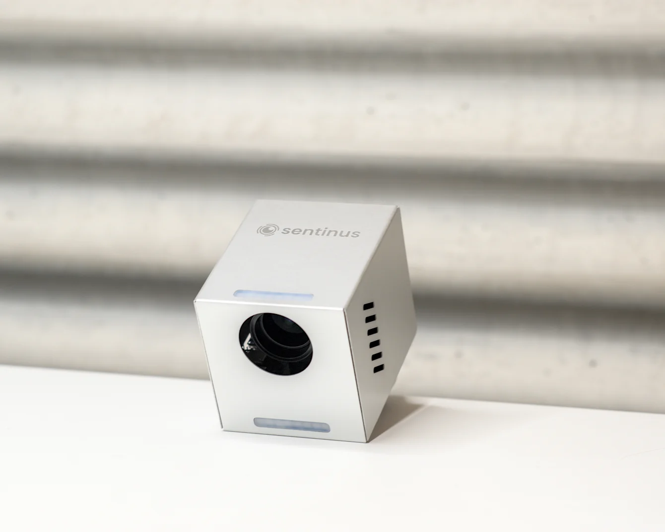 Dispositivo a forma di cubo argentato con una lente, etichettato "sentinus," su una superficie bianca contro uno sfondo grigio testurizzato.