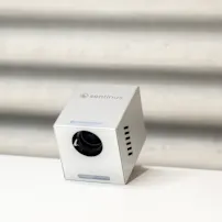 Sølvfarvet kubeformet enhed med en linse, mærket "sentinus," på en hvid overflade mod en tekstureret grå baggrund.