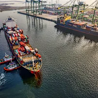 Tàu chở hàng với các container đầy màu sắc neo đậu tại cảng, được hỗ trợ bởi tàu lai, với cần cẩu và container xếp chồng ở phía sau.