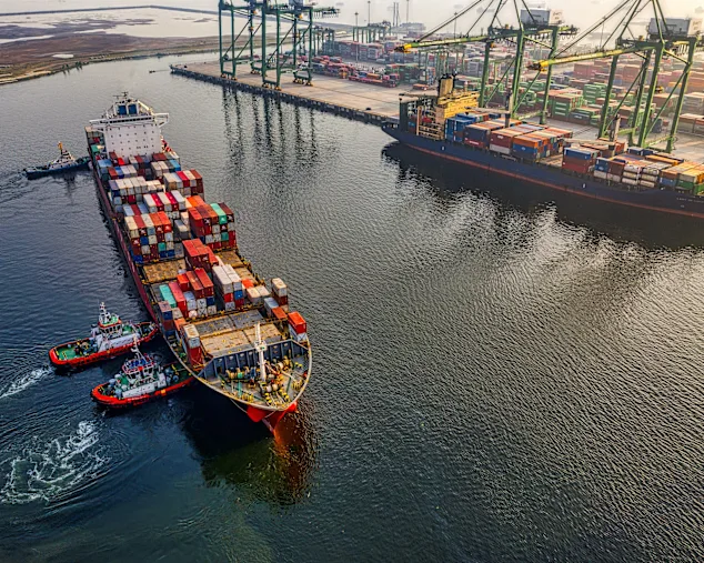 Barco de carga con contenedores coloridos atracado en un puerto, asistido por remolcadores, con grúas y contenedores apilados al fondo.