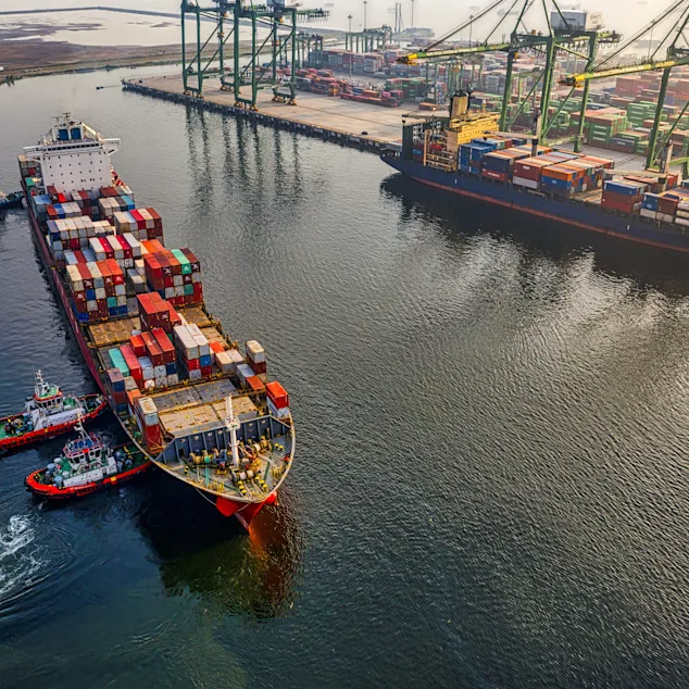 Cargo ship with colorful containers docked at a port, assisted by tugboats, with cranes and stacked containers in the background.