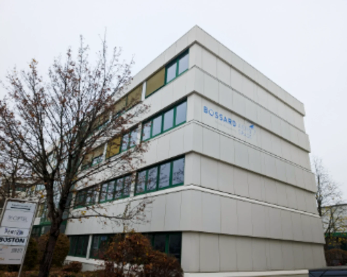 Modern white four-story office building with Bossard Aerospace logo and horizontal windows and bare tree in foreground under overcast sky.