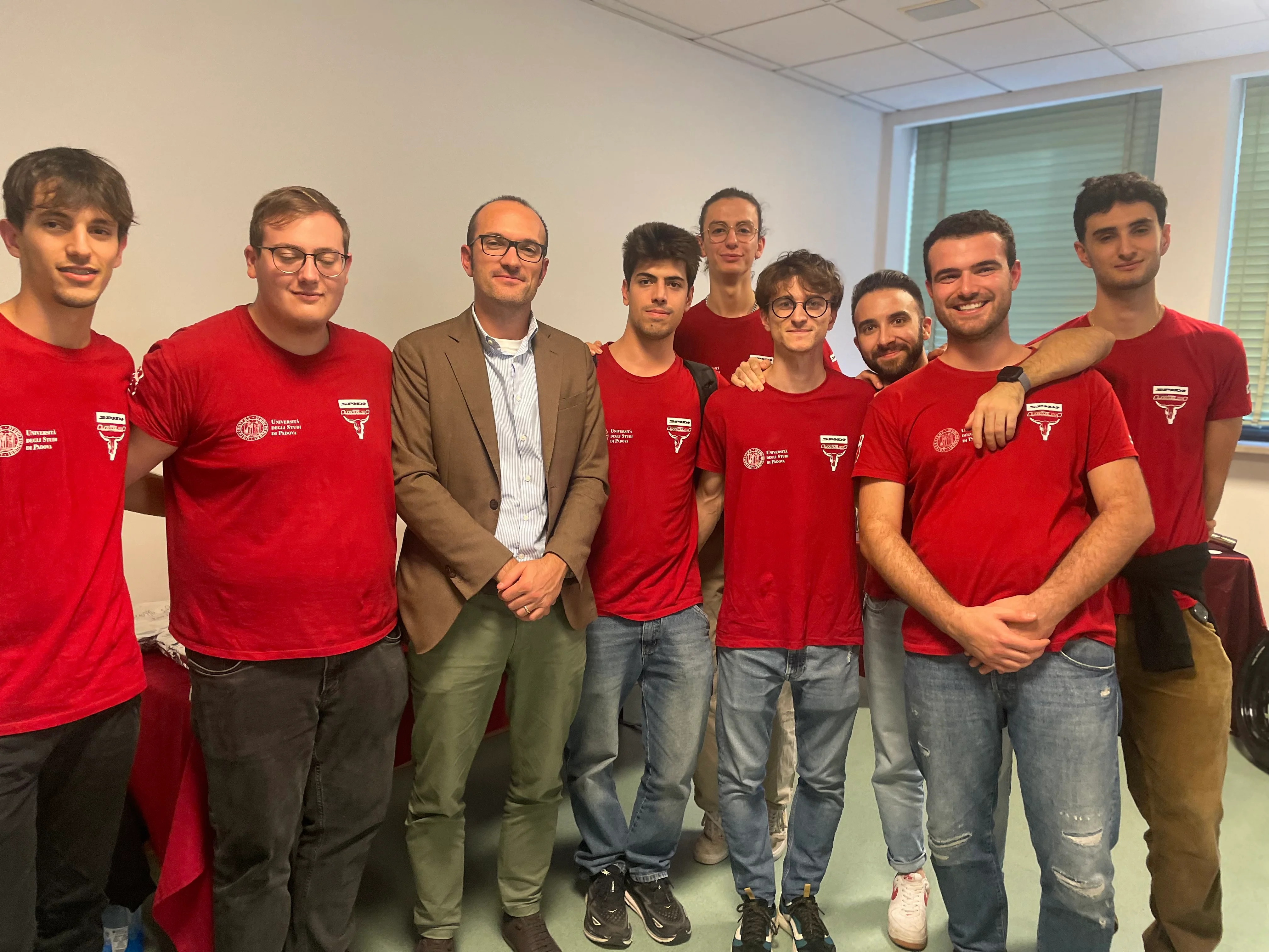 Un gruppo di otto persone, sette con magliette rosse e uno con una giacca marrone, sorride in una stanza con una finestra.