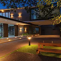 Moderne Hausfassade in der Dämmerung mit warmer Beleuchtung, großen Fenstern und einer Holzbank auf einer Rasenfläche, umgeben von Bäumen.
