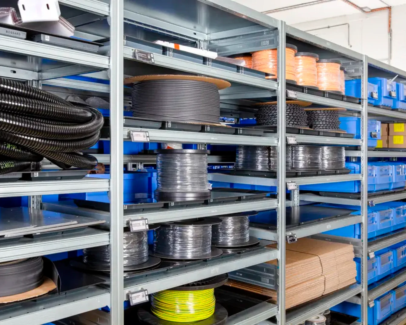 Industrial warehouse shelves with large cable reels, tubing, and blue storage bins organized on metal racks.