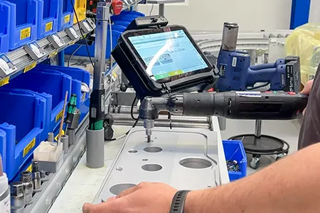 Person, der bruger et elværktøj på en metalplade, med en tablet, der viser instruktioner i nærheden, omgivet af blå kasser.