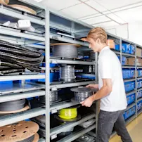 Worker in white shirt retrieving black cable spool from metal shelving unit in organized warehouse.