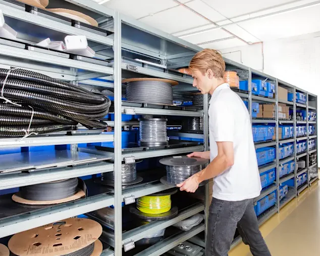 Worker in white shirt retrieving black cable spool from metal shelving unit in organized warehouse.
