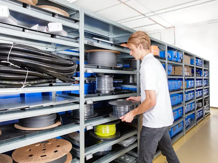 Worker in white shirt retrieving black cable spool from metal shelving unit in organized warehouse.
