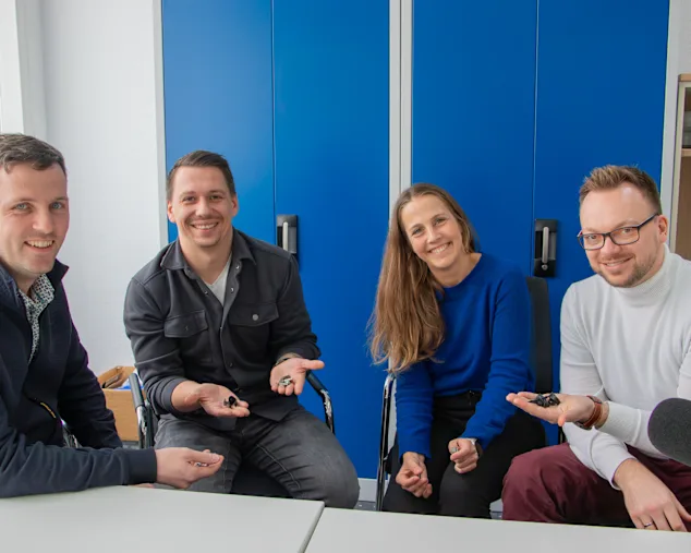 Manuel Aubele, Lukas Holitschke, Maja Matzke and Tobias Engelberg sitzen nebeneinander vor einem blauen Aktenschrank. Vor ihnen steht ein schwarzes Studiomikrofon, sie halten Clip Befestiger in den Händen und lächeln in die Kamera