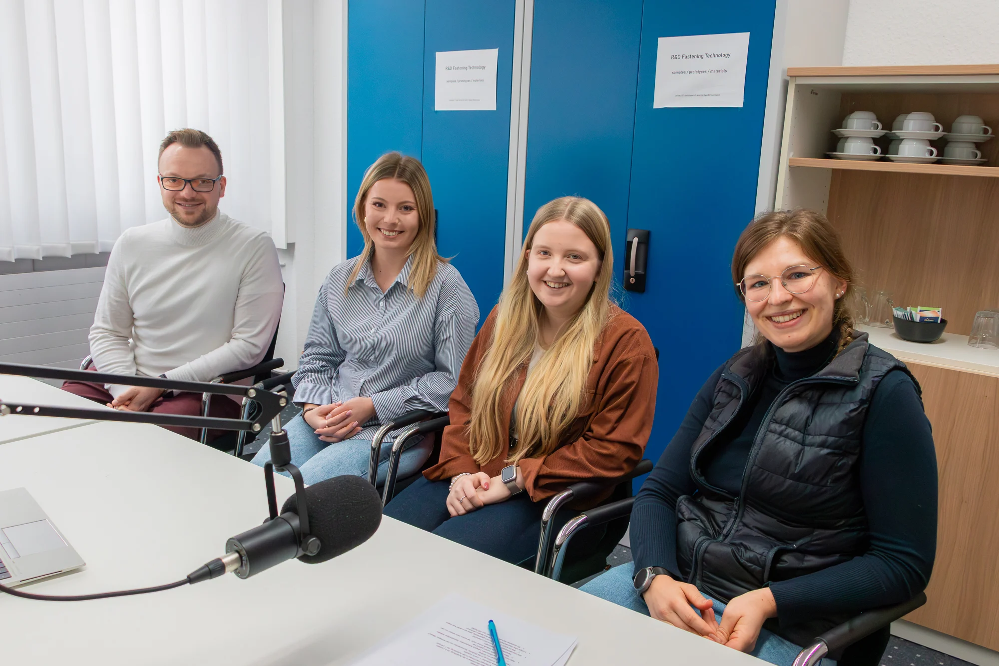 Tobias Engelberg, Alina Iwich, Alexa Häger und Raffaela Mikulla sitzen nebeneinander vor einem Broadcast Mikrofon und lächeln in die Kamera. Im Hintergrund ist blauer Aktenschrank und ein braunes Regal mit Tassen.