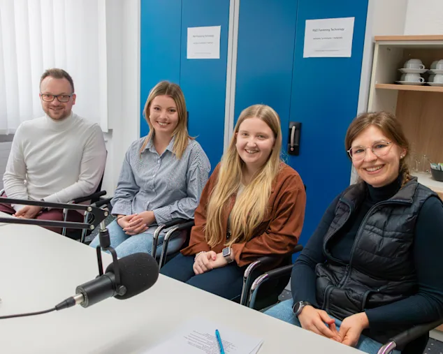 Tobias Engelberg, Alina Iwich, Alexa Häger und Raffaela Mikulla sitzen nebeneinander vor einem Broadcast Mikrofon und lächeln in die Kamera. Im Hintergrund ist blauer Aktenschrank und ein braunes Regal mit Tassen.