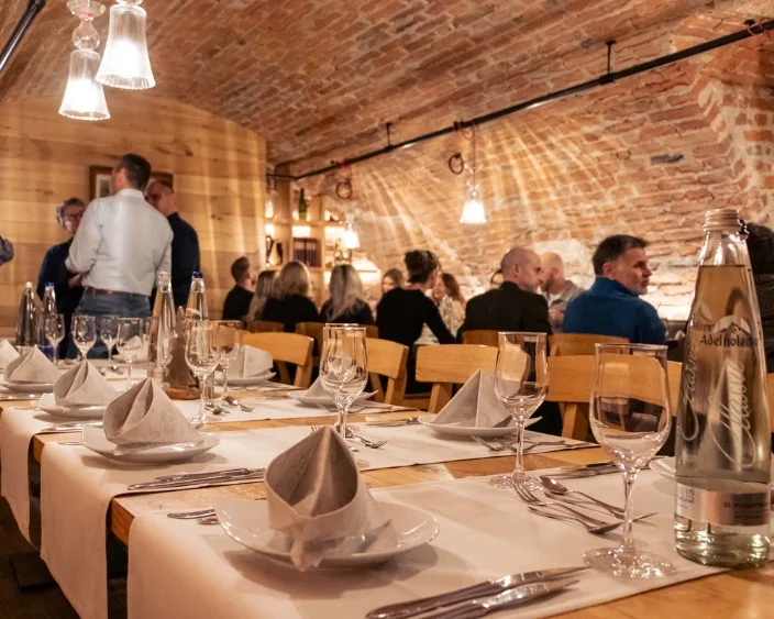 Gedeckter Tisch in einem Gewölbekeller mit weißen Servietten, Gläsern und Besteck. Im Hintergrund sitzen und stehen mehrere Personen in Gruppen zusammen und unterhalten sich.