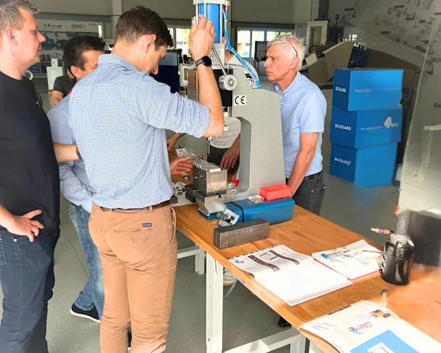 Skupina mužů v Bossard Experience Center v Linci zkoumá bílý a modrý hydraulický lis na stole s technickou dokumentací.
