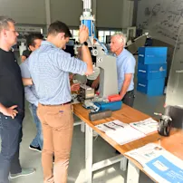 Skupina mužů v Bossard Experience Center v Linci zkoumá bílý a modrý hydraulický lis na stole s technickou dokumentací.