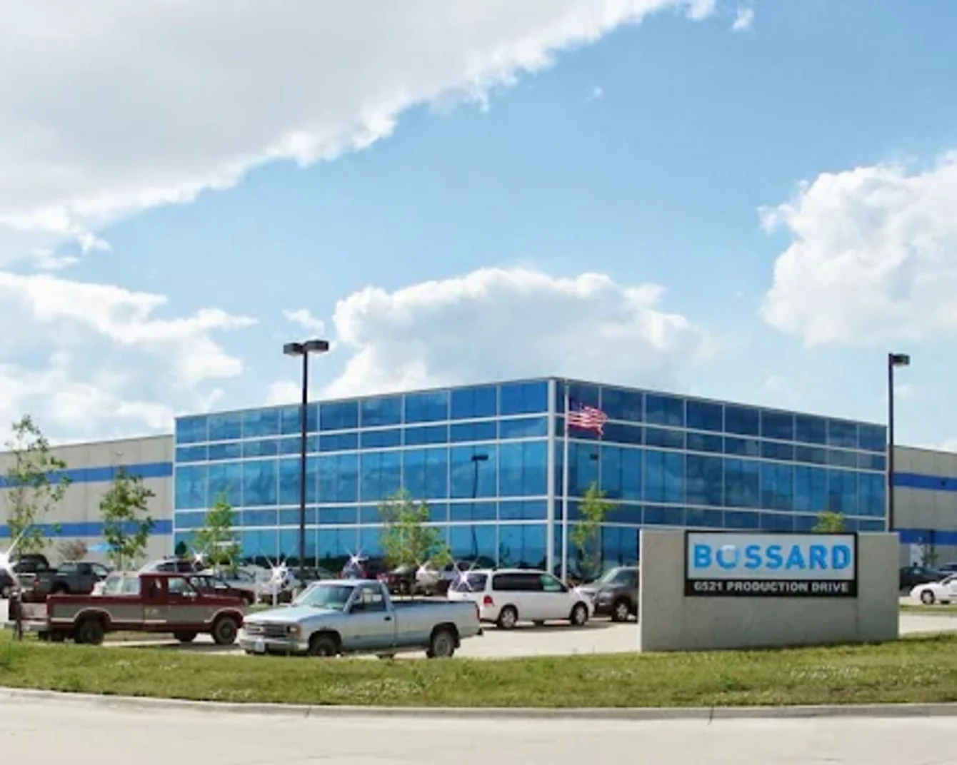 Large industrial building with blue-tinted windows, surrounded by parked cars and a sign reading "Bossard." Cloudy sky above.