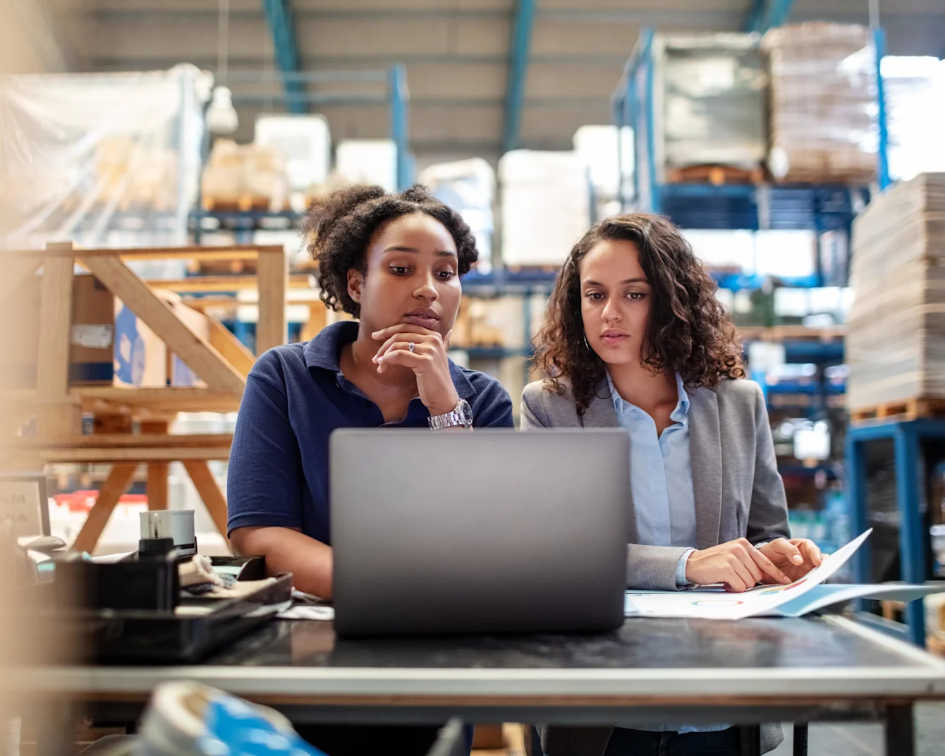 Two women reviewing laptop and documents at desk in warehouse with blue shelving and stacked inventory in background.