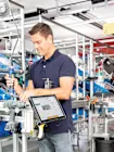 Man in een blauw shirt werkt met gereedschap aan een assemblagelijn, omringd door blauwe bakken en een tablet die informatie weergeeft.