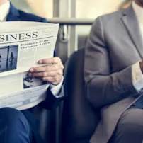 A man reads a newspaper while another man next to him reads on his cell phone