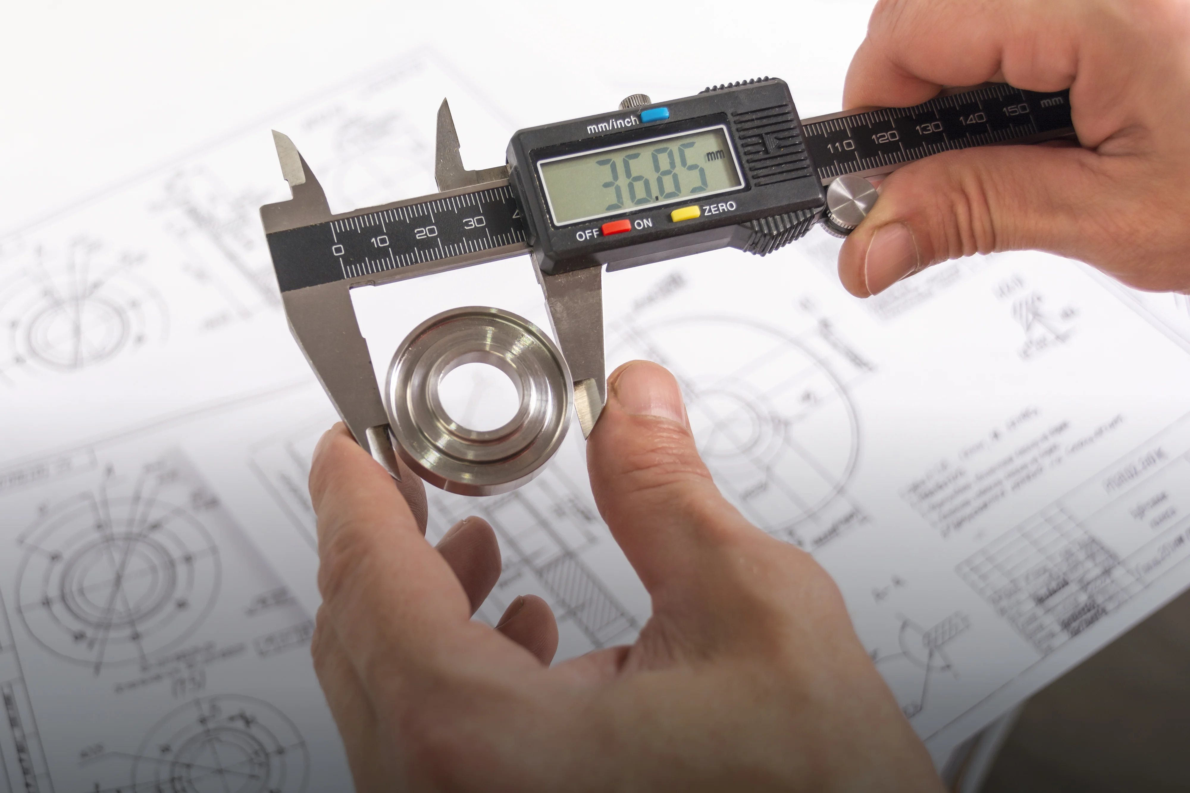 A pair of hands gripping a caliper to measure a metal bearing above technical drawings and specifications.