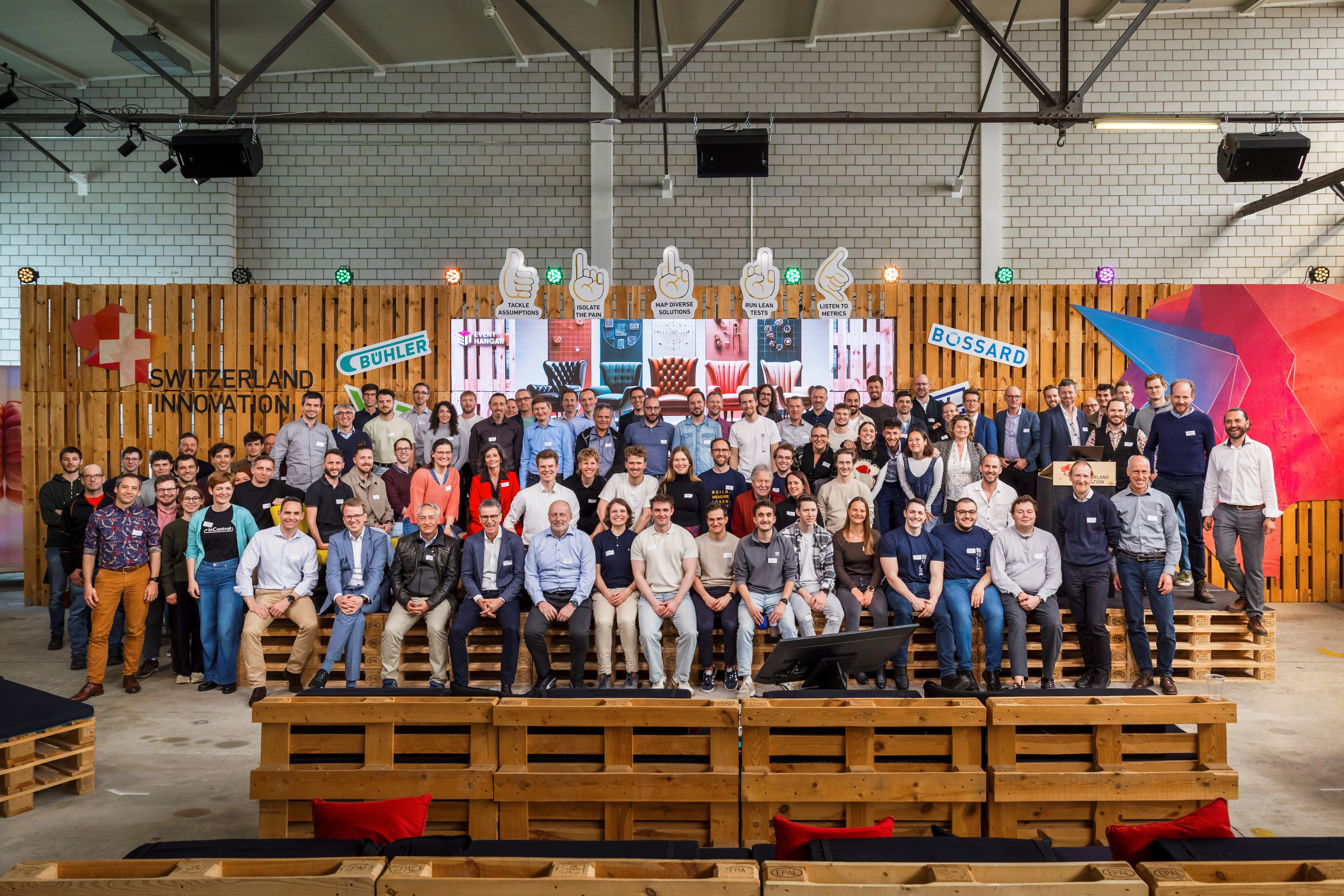 Un grand groupe de personnes pose sur des bancs en bois dans un entrepôt, avec "Switzerland Innovation" et des logos en arrière-plan.