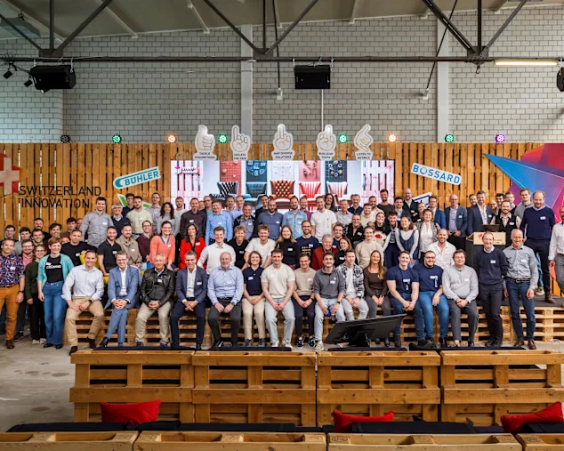 A large group of people pose on wooden benches in a warehouse setting, with "Switzerland Innovation" and logos in the background.