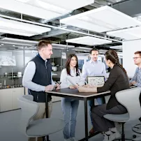 Persone intorno a un tavolo discutono di un disegno tecnico in un ufficio dall'aspetto industriale.