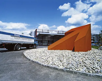 Historical image of a large orange arrow sculpture in a roundabout, with a Bossard truck and warehouse in the background.