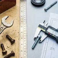 A top-down view of an engineering workspace featuring an electronic digital caliper measuring the thread diameter of a bolt