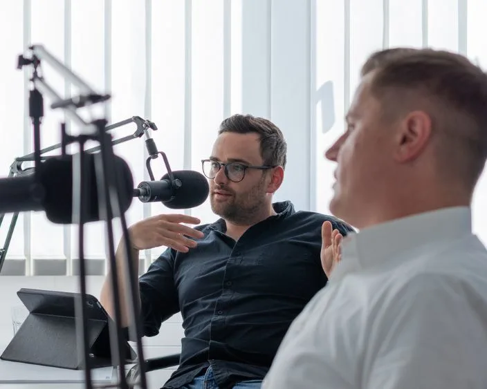 Zwei Experten sitzen in einem hellen weißen Raum vor Broadcast Mikrofonen, der hintere Gast spricht gestikulierend, ein Tablet steht auf dem Tisch.