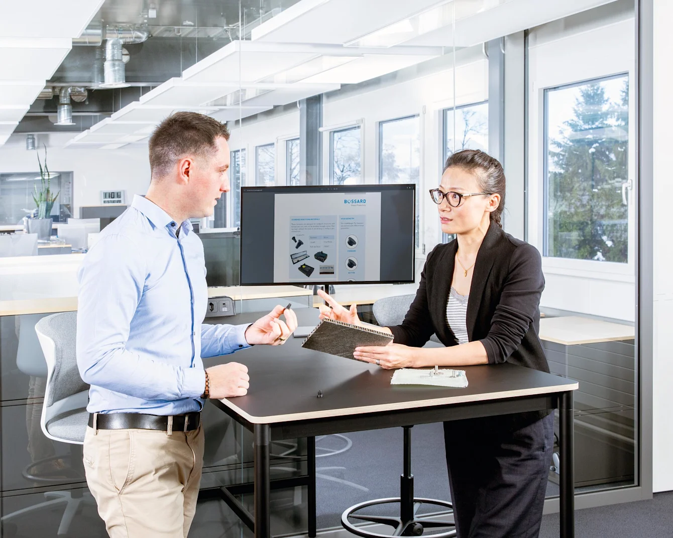 Two people at a desk in a modern office discuss fastening a composite material; one holds a sandwich panel, the other a fastener.