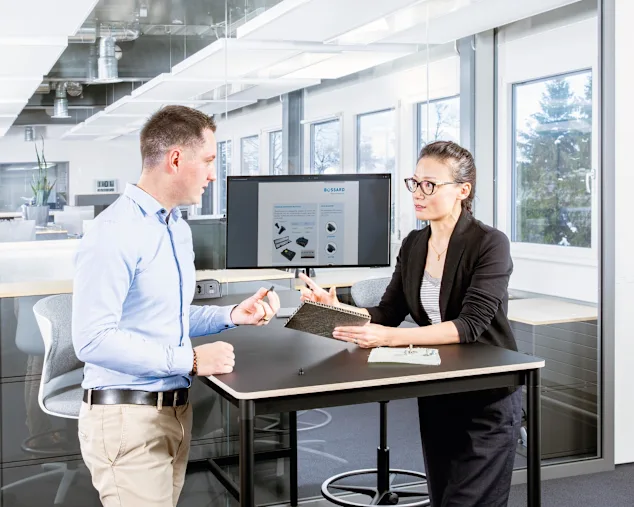 Two people at a desk in a modern office discuss fastening a composite material; one holds a sandwich panel, the other a fastener.