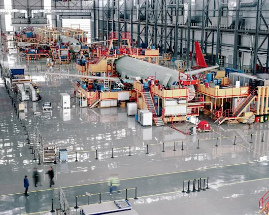 Et stort fly bliver samlet inde i en rummelig fabrik, hvor industrielt maskineri og arbejdere ses i baggrunden.
