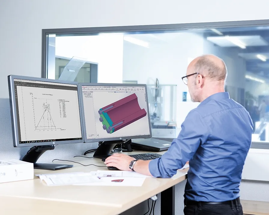 Un uomo con una camicia blu lavora su un disegno tecnico a una scrivania in piedi al computer.