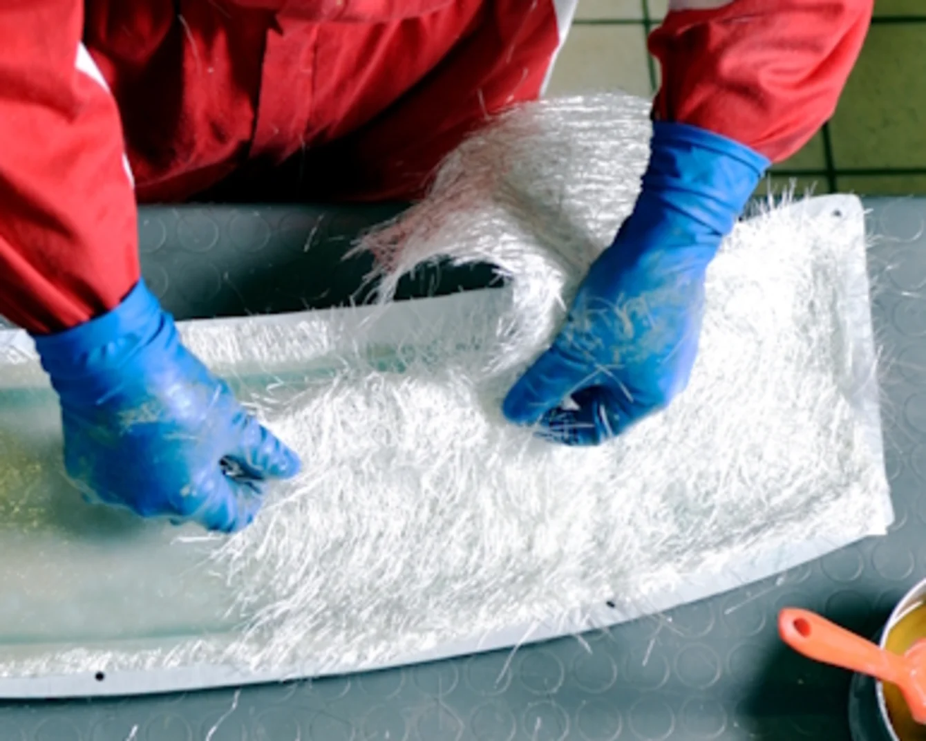 Person in roten Arbeitskleidung und blauen Handschuhen verarbeitet Glasfasermatten auf einer Form.